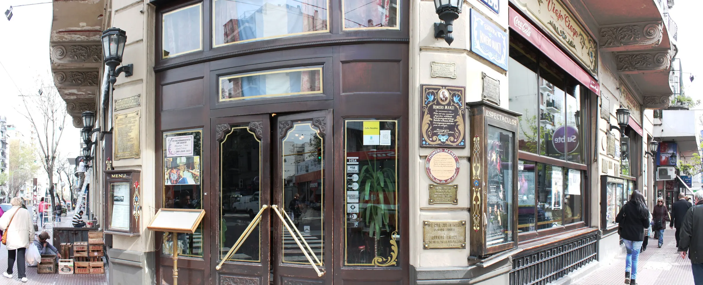 L'ingresso curvo d'angolo dell'Esquina Homero Manzi all'incrocio tra San Juan e Boedo, con porte in legno scuro, maniglie in ottone, targhe in ottone e insegne dipinte sulle pareti ai due lati