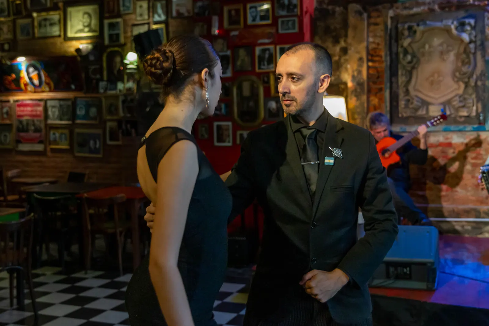 Una coppia di tango vestita di nero che balla in un bar in Argentina mentre un chitarrista suona dietro di loro, fotografata in una sede diversa